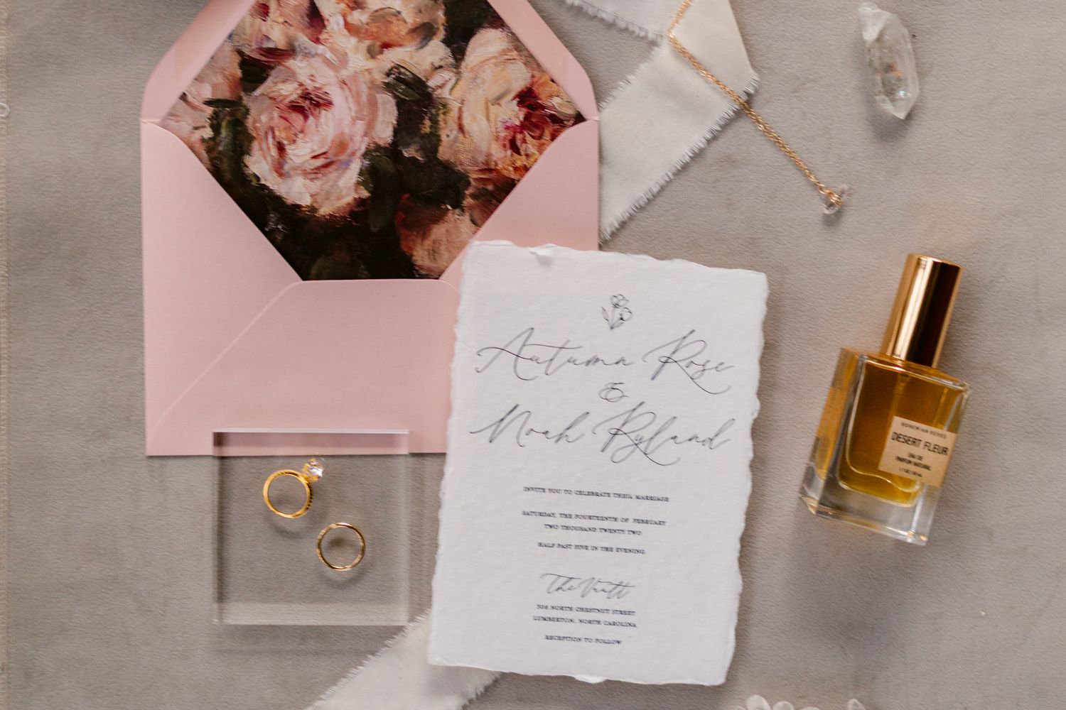 Elegant wedding stationery flatlay with pink roses, perfume bottle, and delicate calligraphy on light grey background.