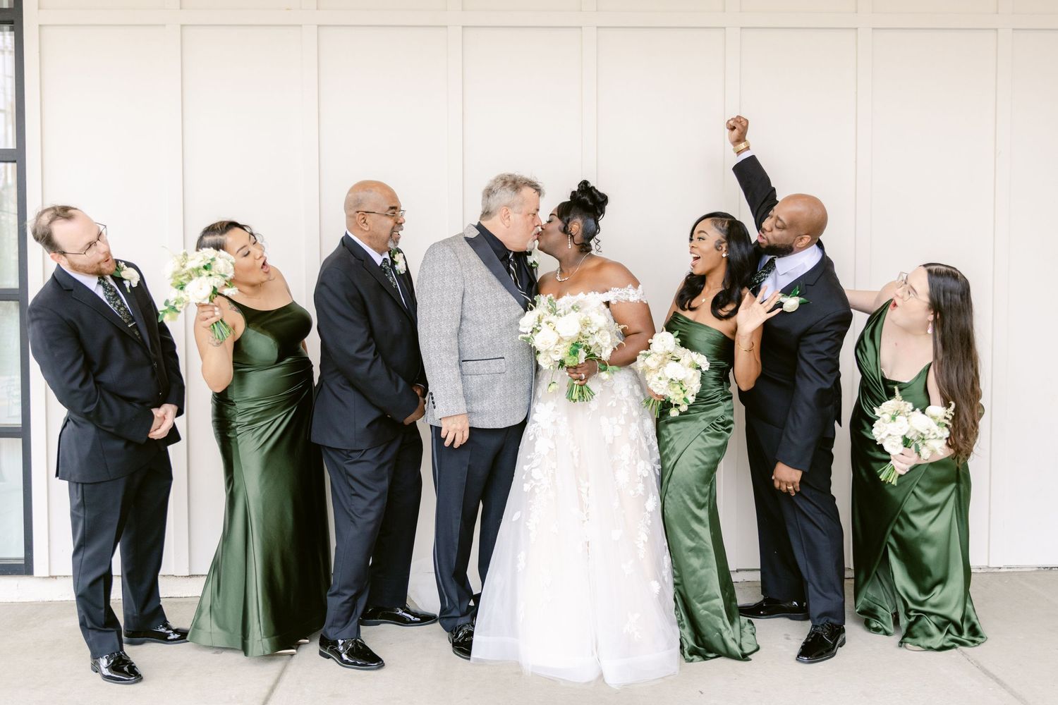 Wedding party in green dresses and black suits pose playfully in a line against a white wall.