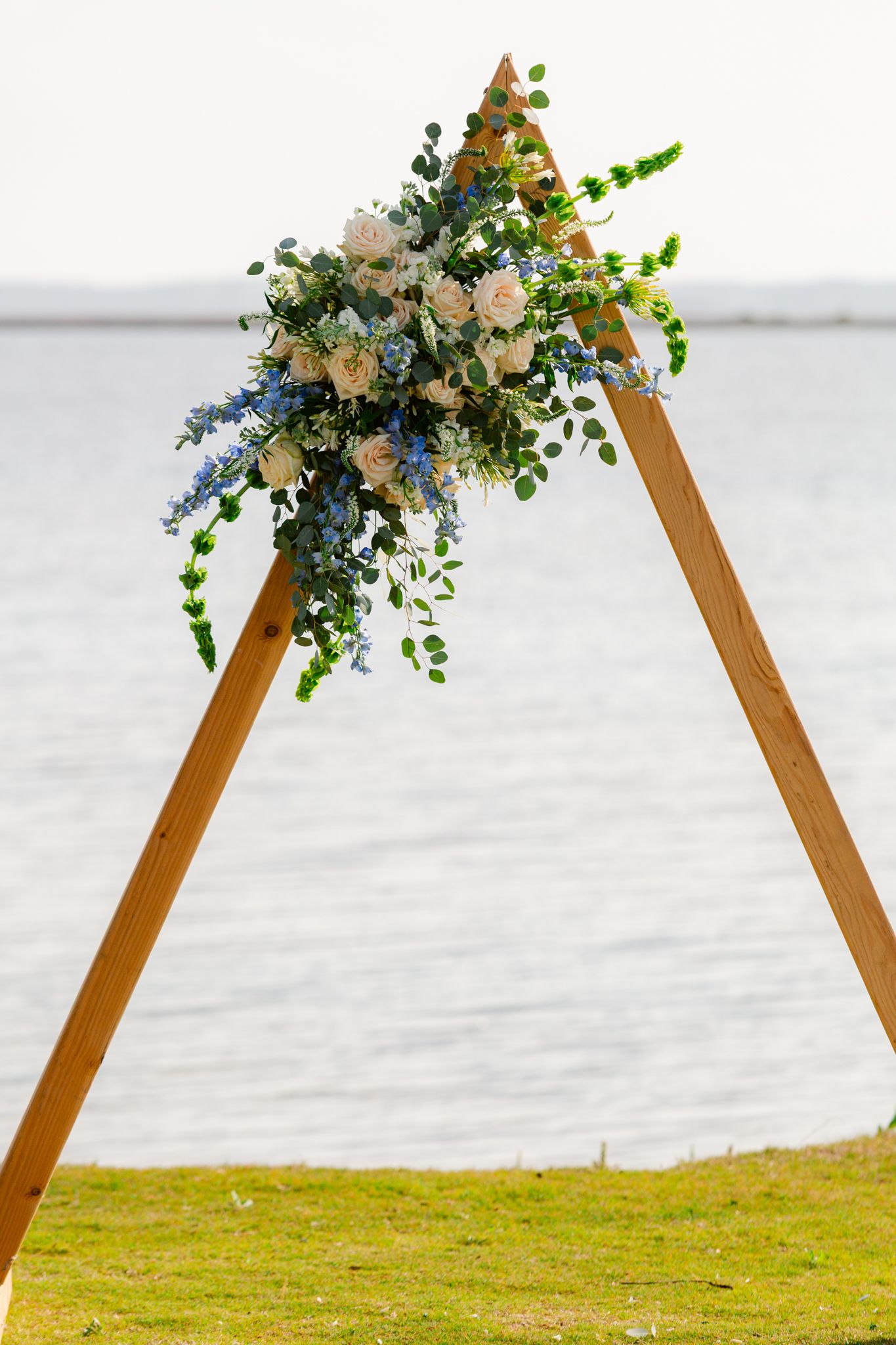 Wooden wedding arch decorated with lush white flowers and greenery stands on grass overlooking a waterfront view.