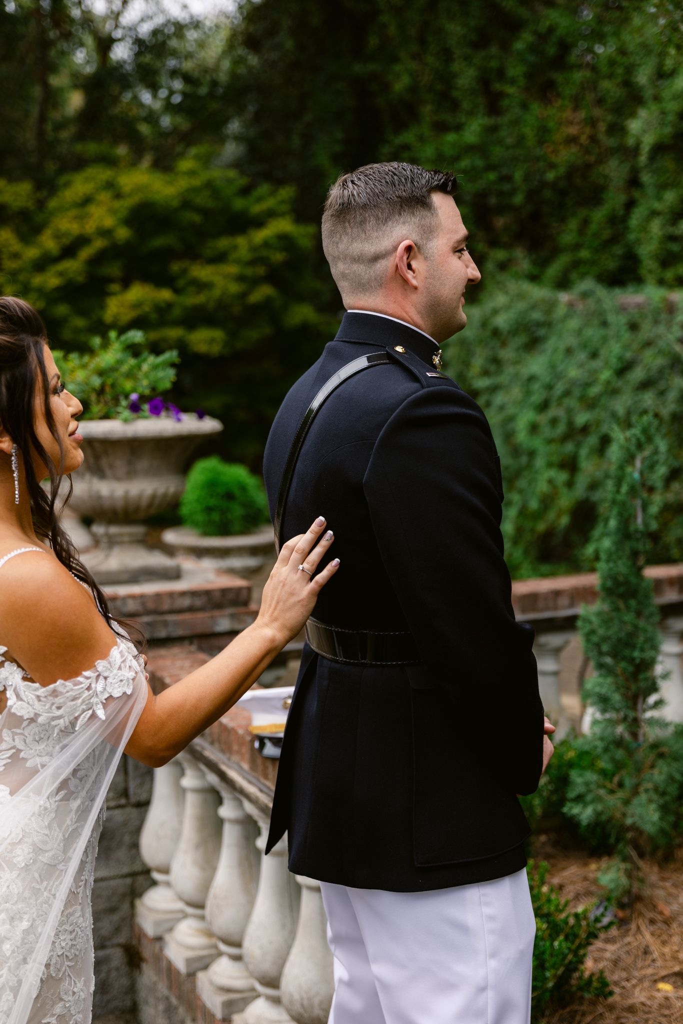A couple in wedding attire shares a playful first look moment in a garden setting with stone balustrades.