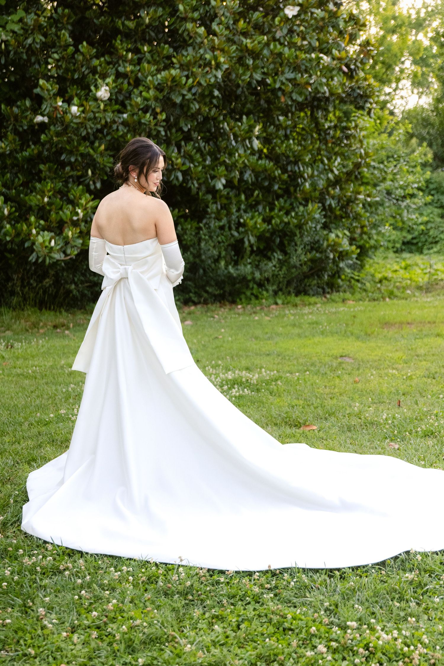 A flowing white wedding dress with a dramatic train cascades across a sunlit garden lawn.