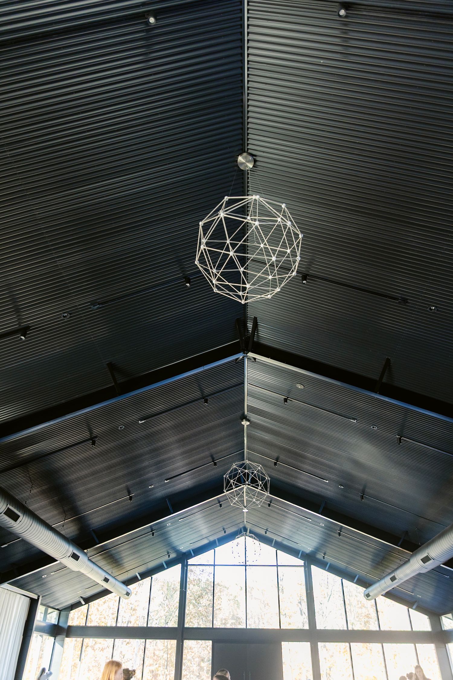 Modern geometric light fixtures hang from a dark vaulted ceiling with large windows in an architectural space.