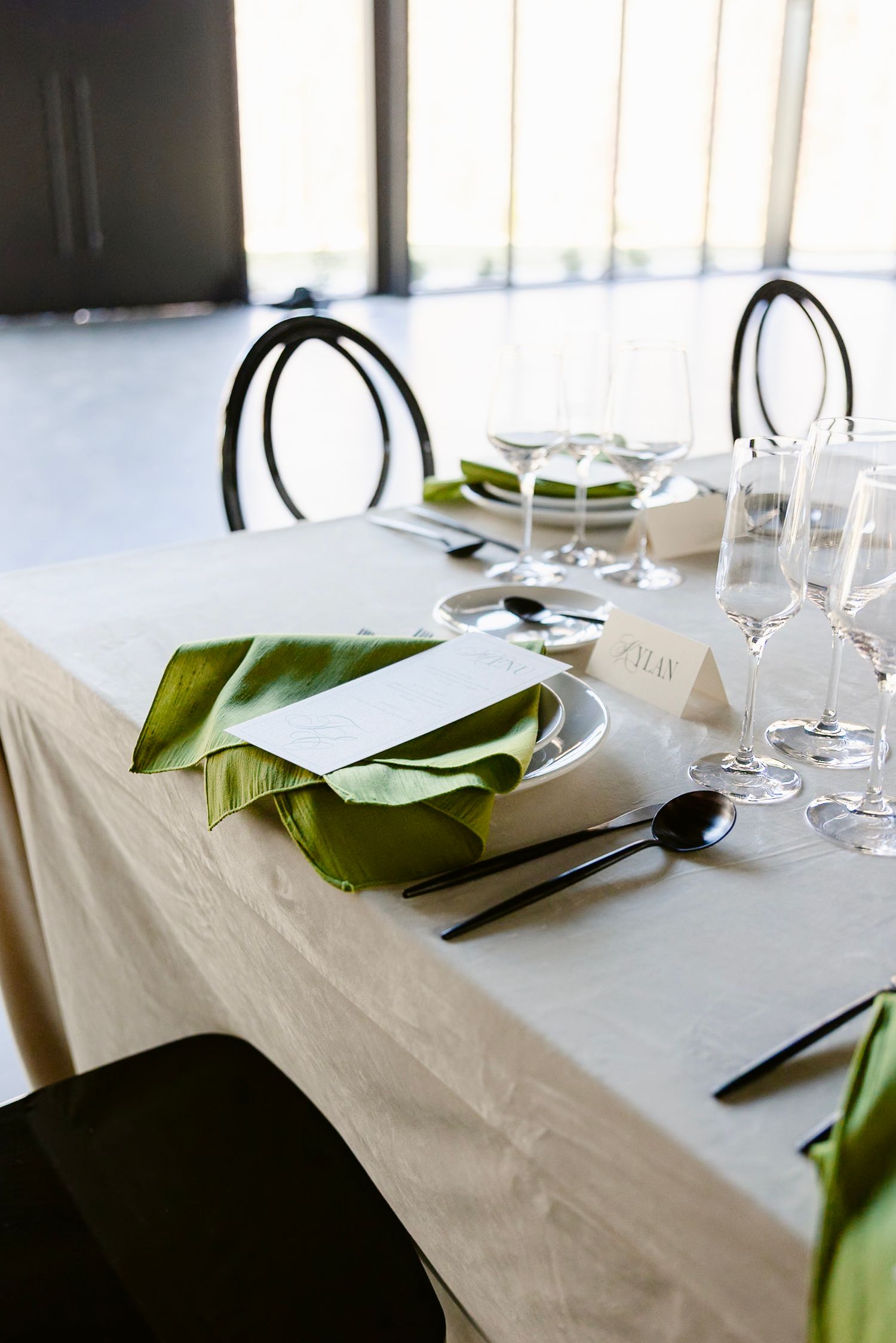 Elegant table setting with white tablecloth, green napkins, wine glasses and black chairs in a modern dining room.