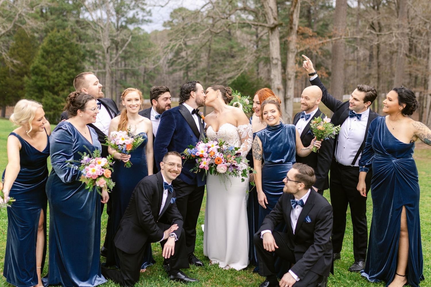 Wedding party in navy blue and white attire laughs together during outdoor photo session in a garden setting.