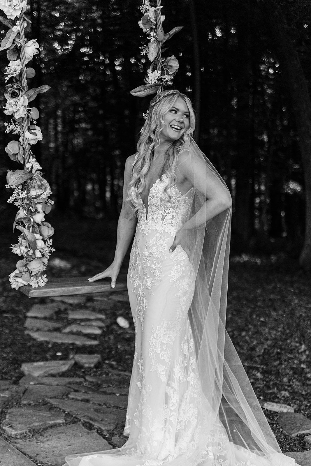 A bride in a delicate lace wedding dress poses outdoors among trees with a flowing cathedral veil.