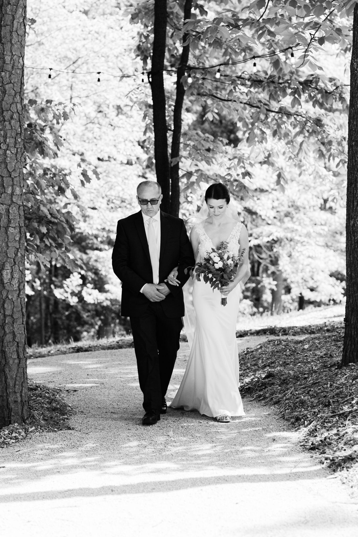 A black and white photo shows a couple walking down a tree-lined path during a wedding ceremony.