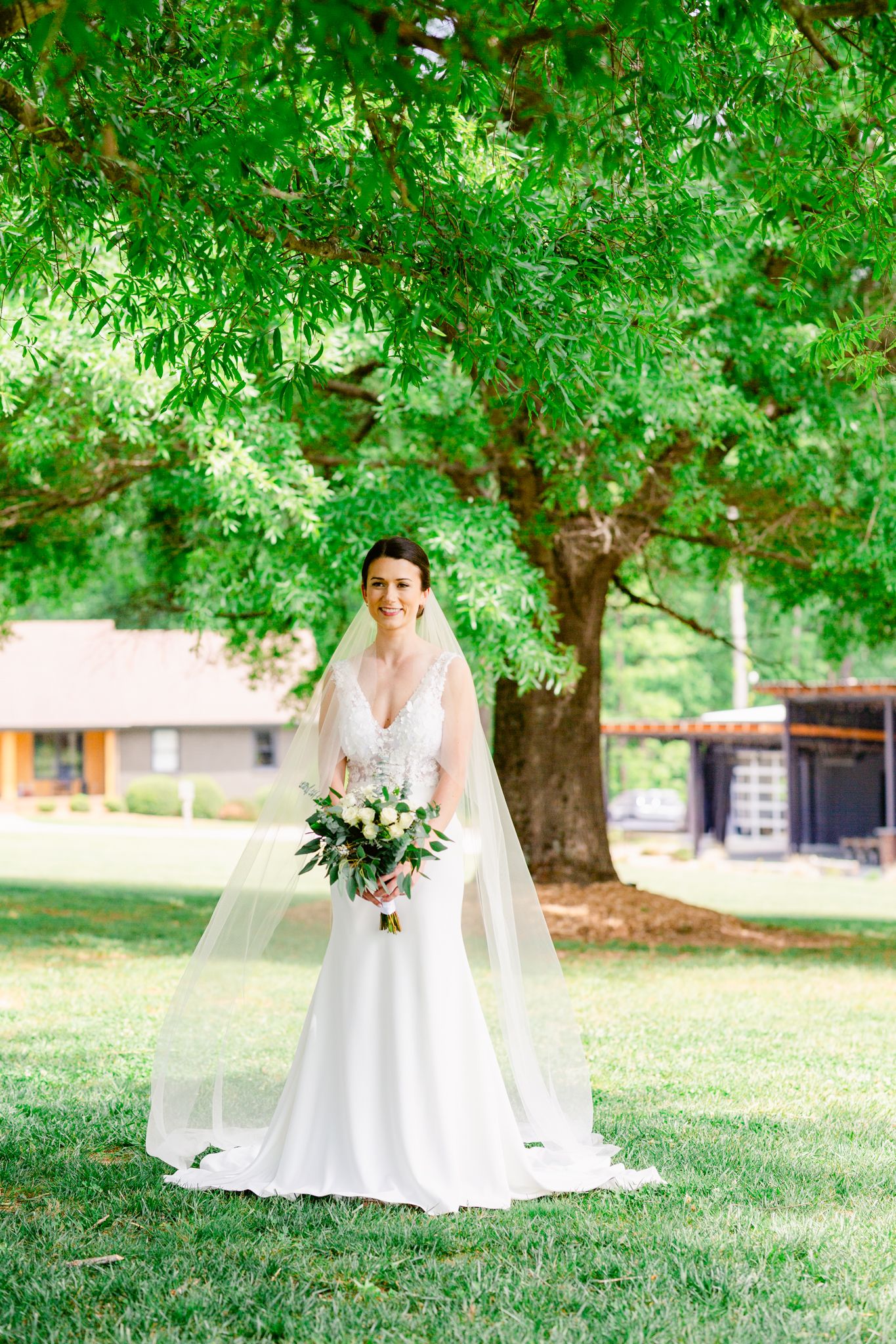 A wedding dress flows elegantly across a lush green lawn beneath the shade of mature trees in a summer garden setting.