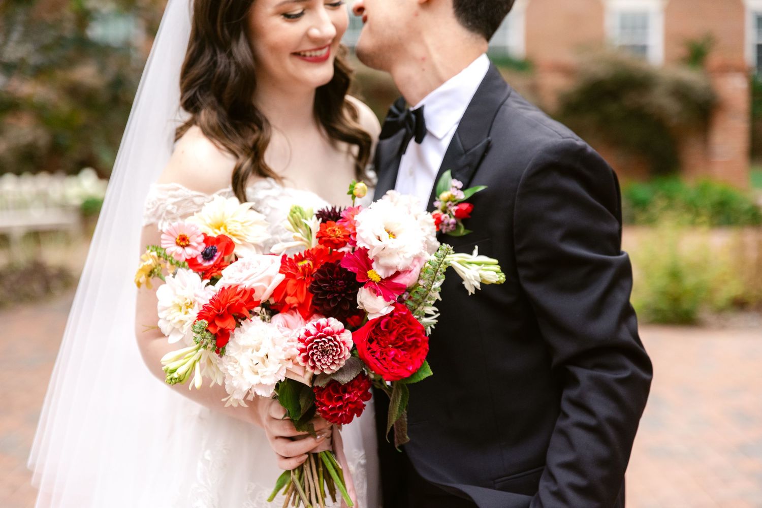 A couple shares an intimate moment outdoors while holding a vibrant red and white wedding bouquet.