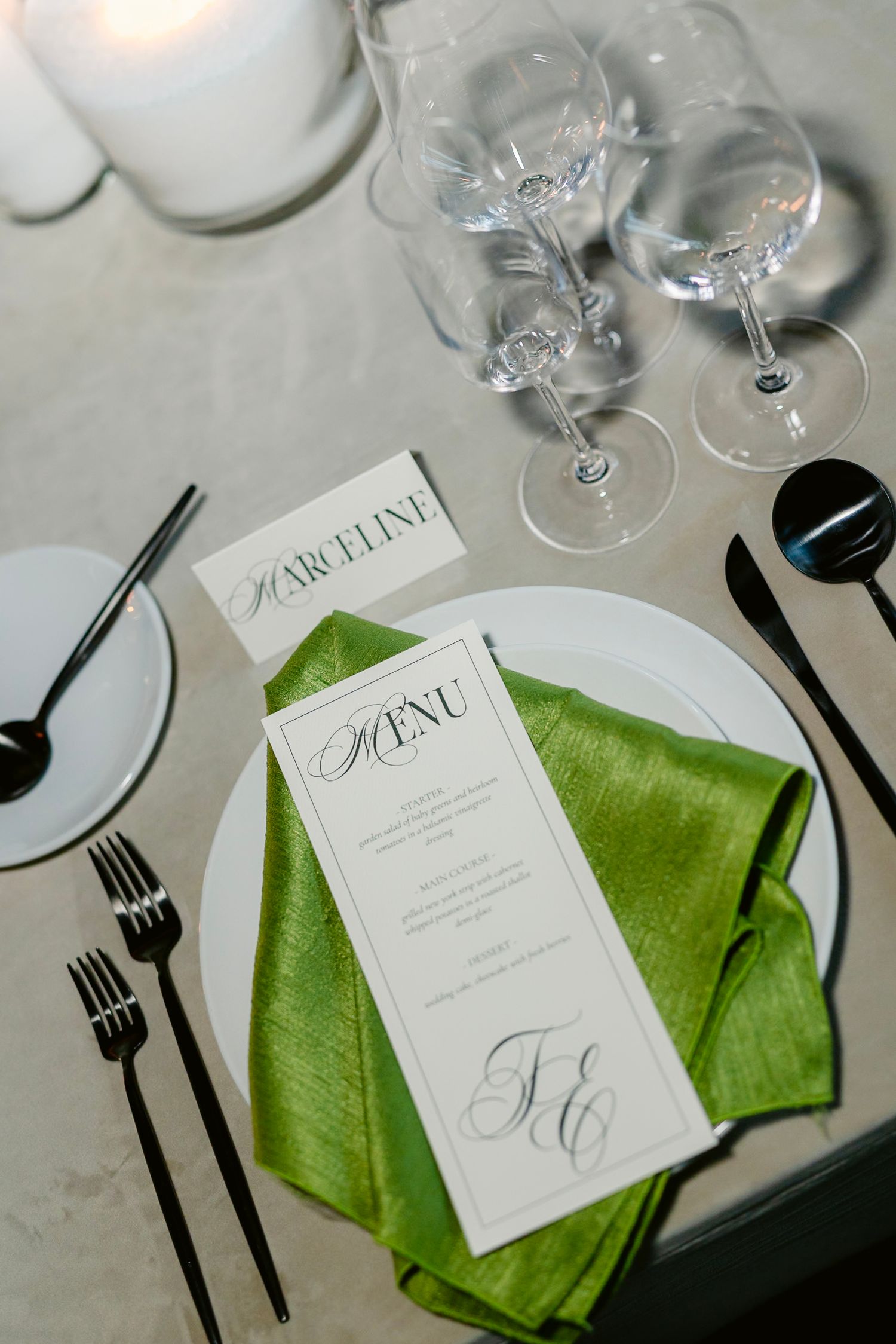 Elegant table setting with menu card on lime green napkin, wine glasses and black cutlery on white plate.