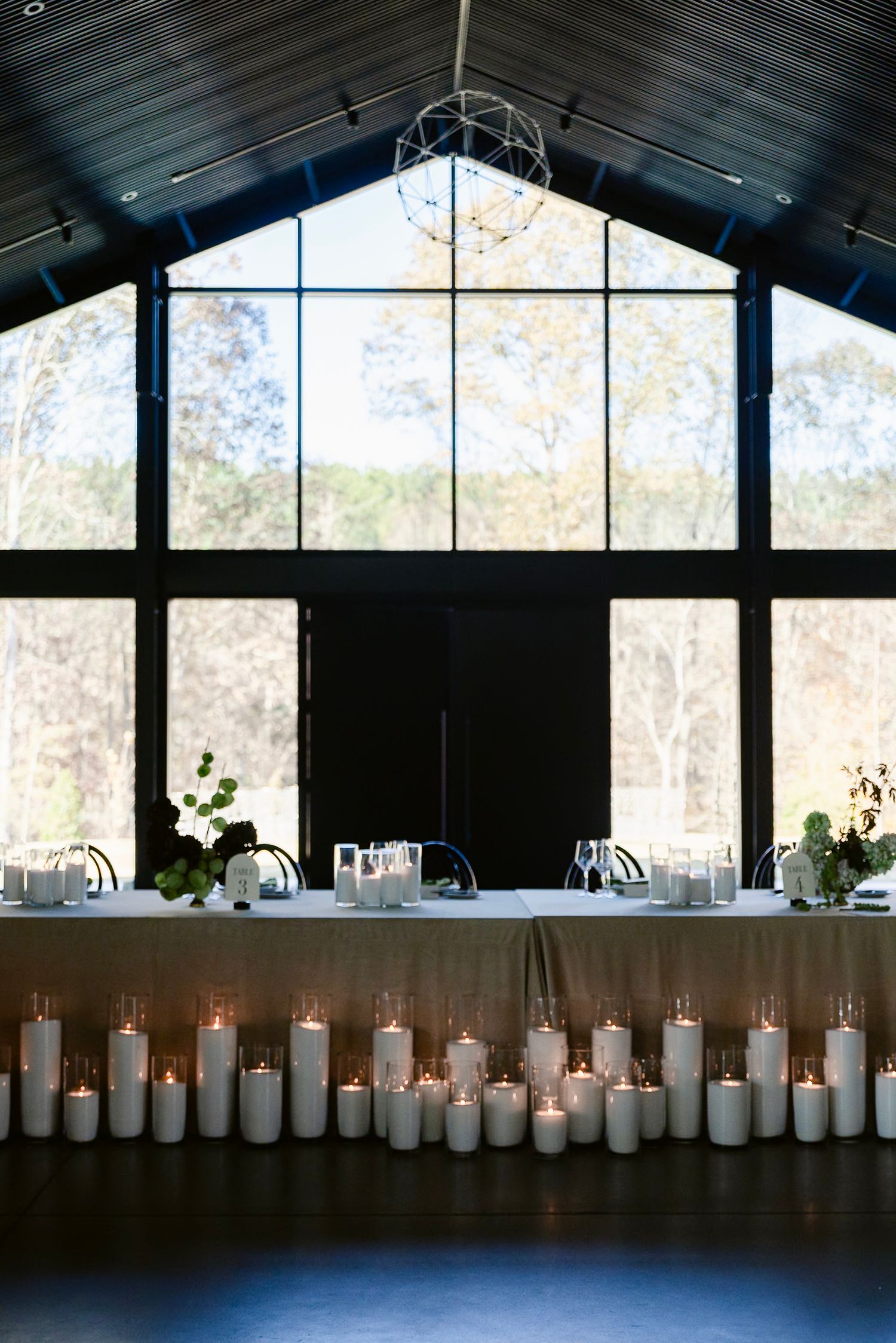 A row of white pillar candles lines a dark wooden table in an elegant venue with large windows and natural light.