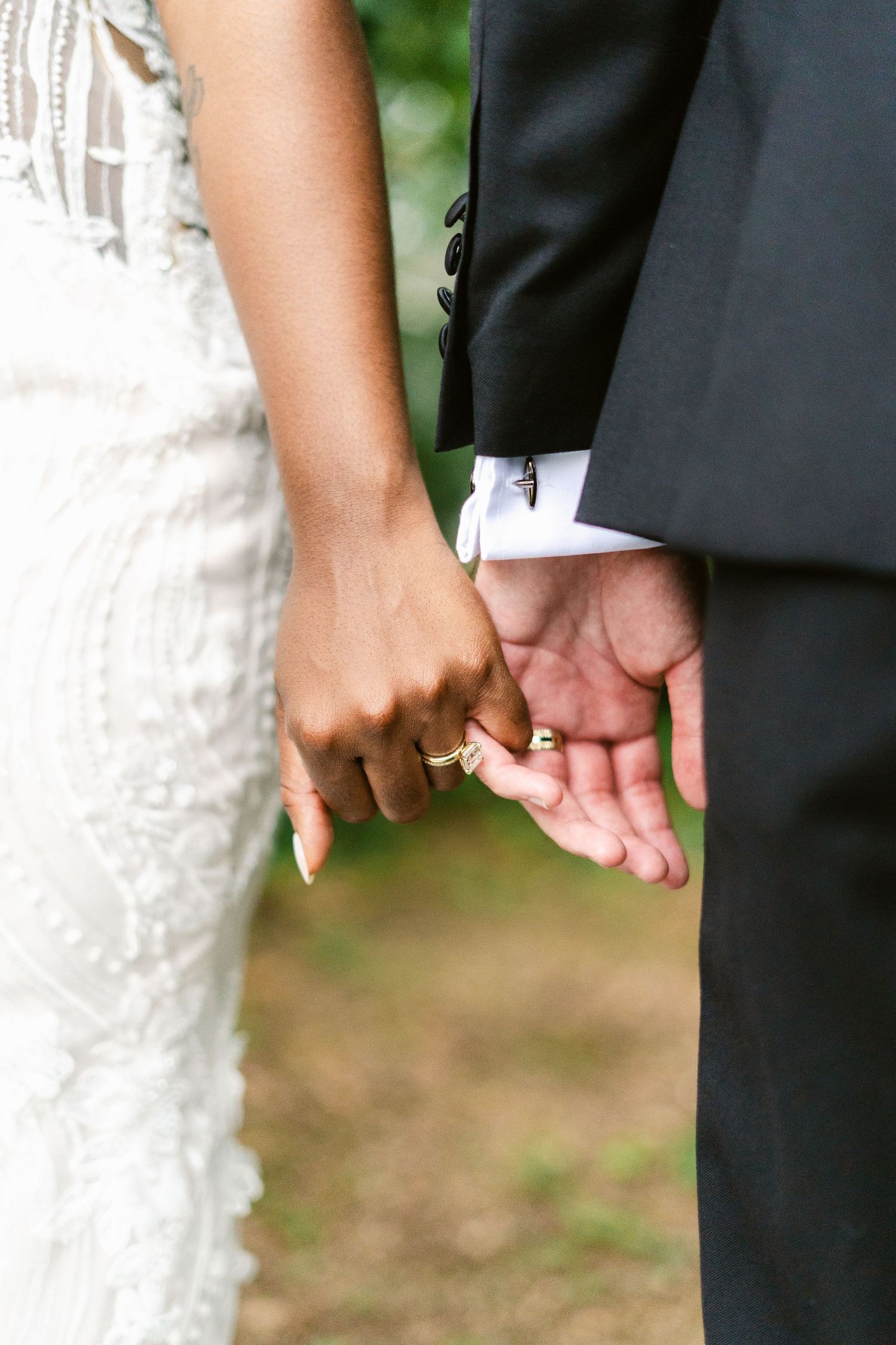 Close-up of newlyweds holding hands, showing white lace dress and black suit details.