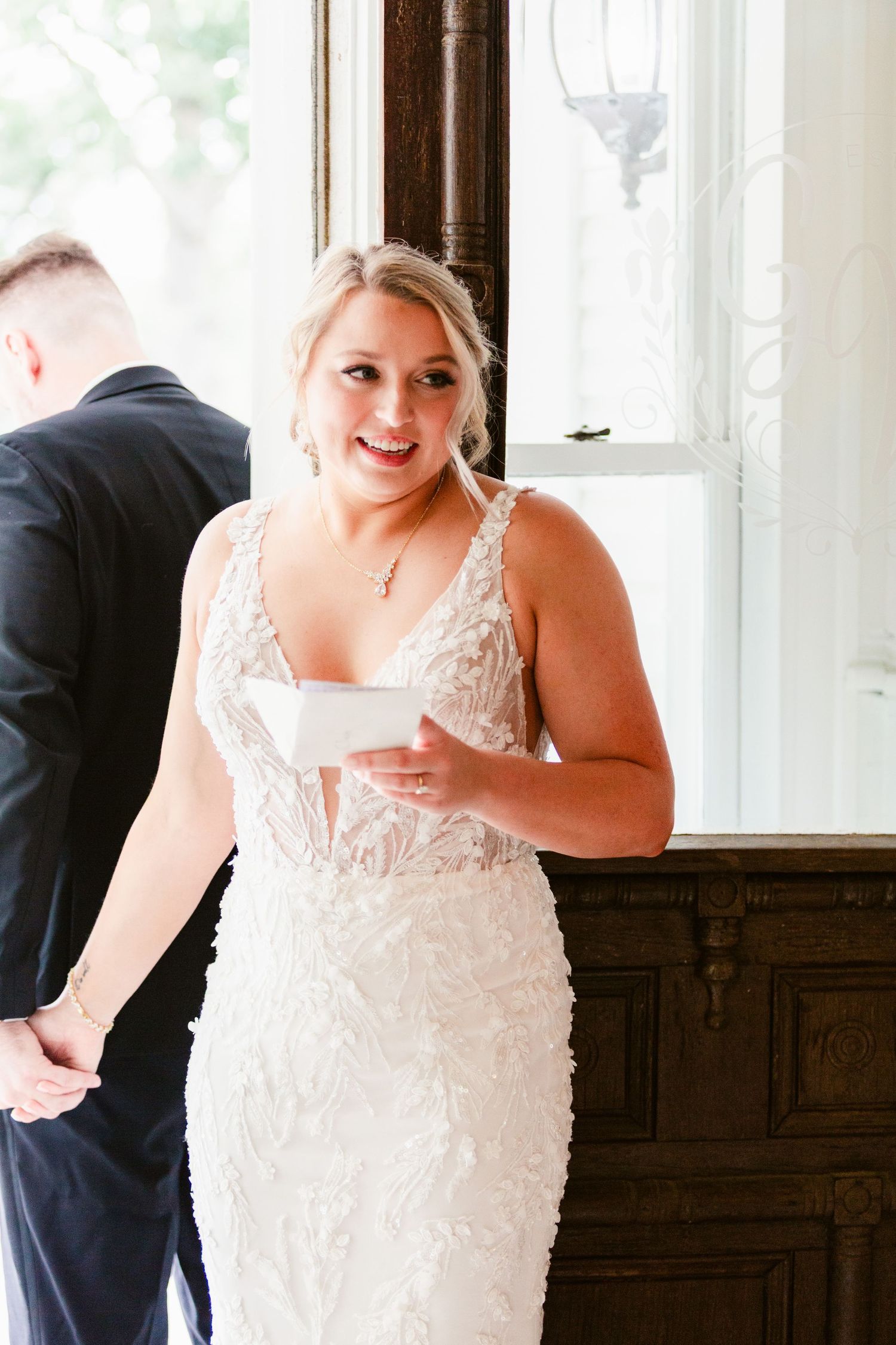 Bride in an elegant white lace wedding gown with delicate straps holds a glass during a reception.