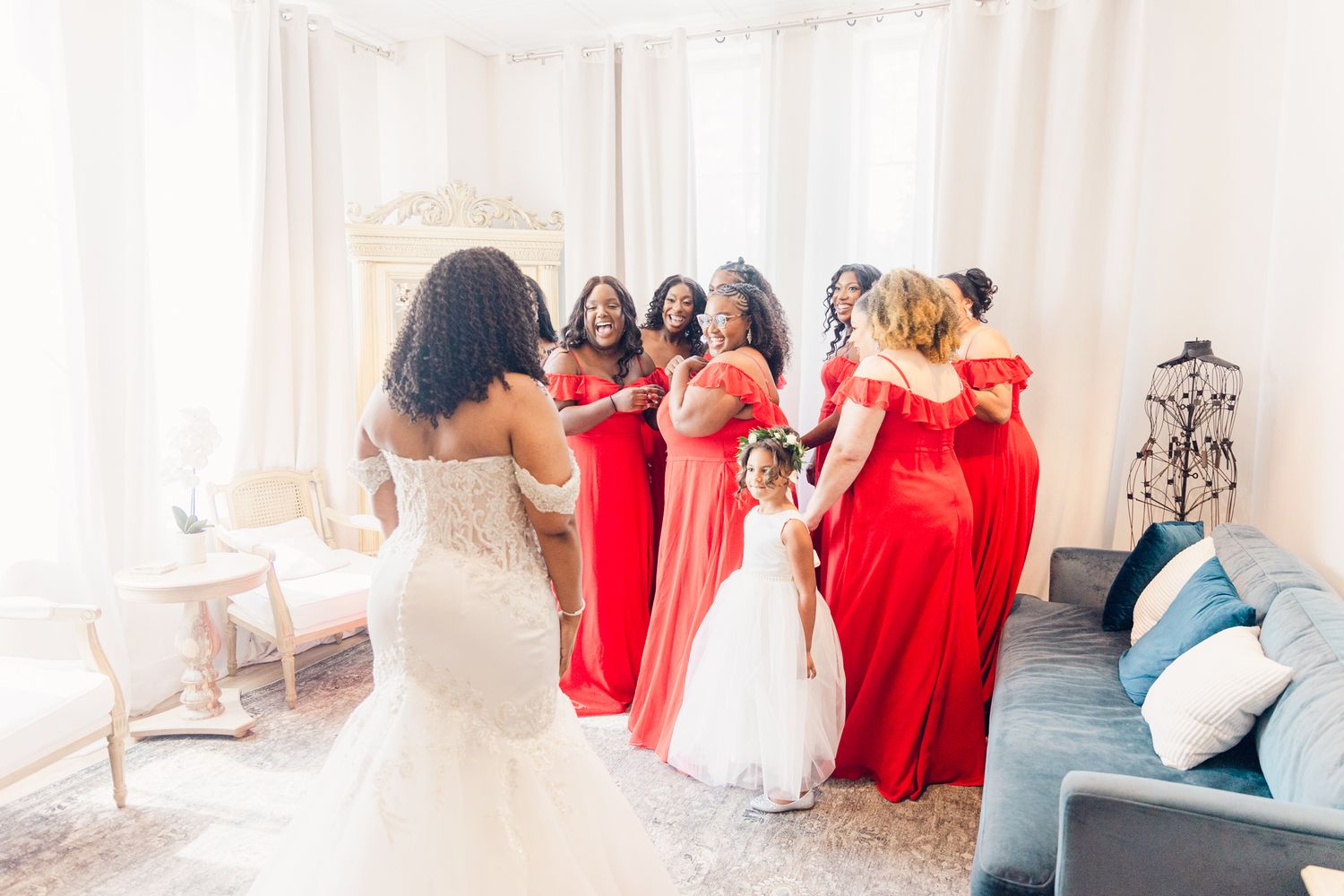 Wedding party members in red dresses and white gown pose together in an elegant light-filled room.