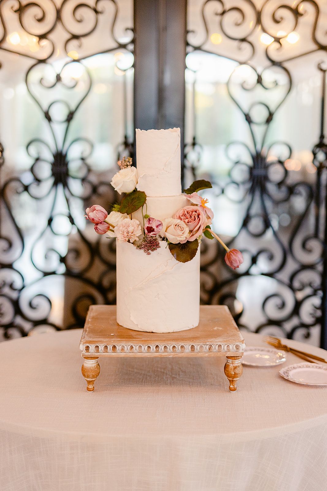 Minimalist white wedding cake adorned with pink flowers on an ornate silver stand against decorative ironwork.