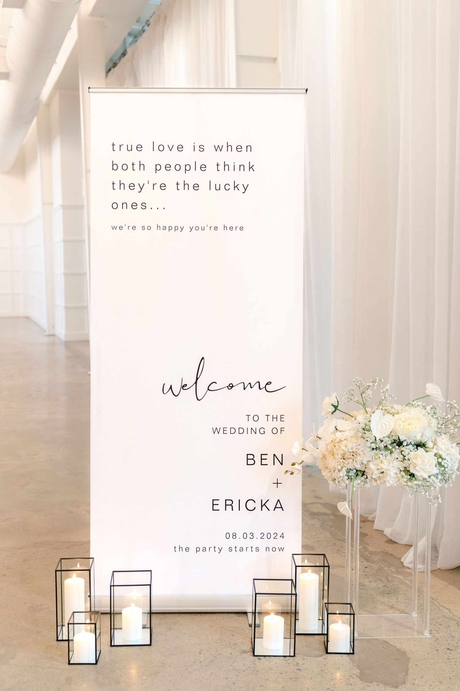 Minimalist wedding welcome sign display with candles and floral arrangements in a bright white venue.