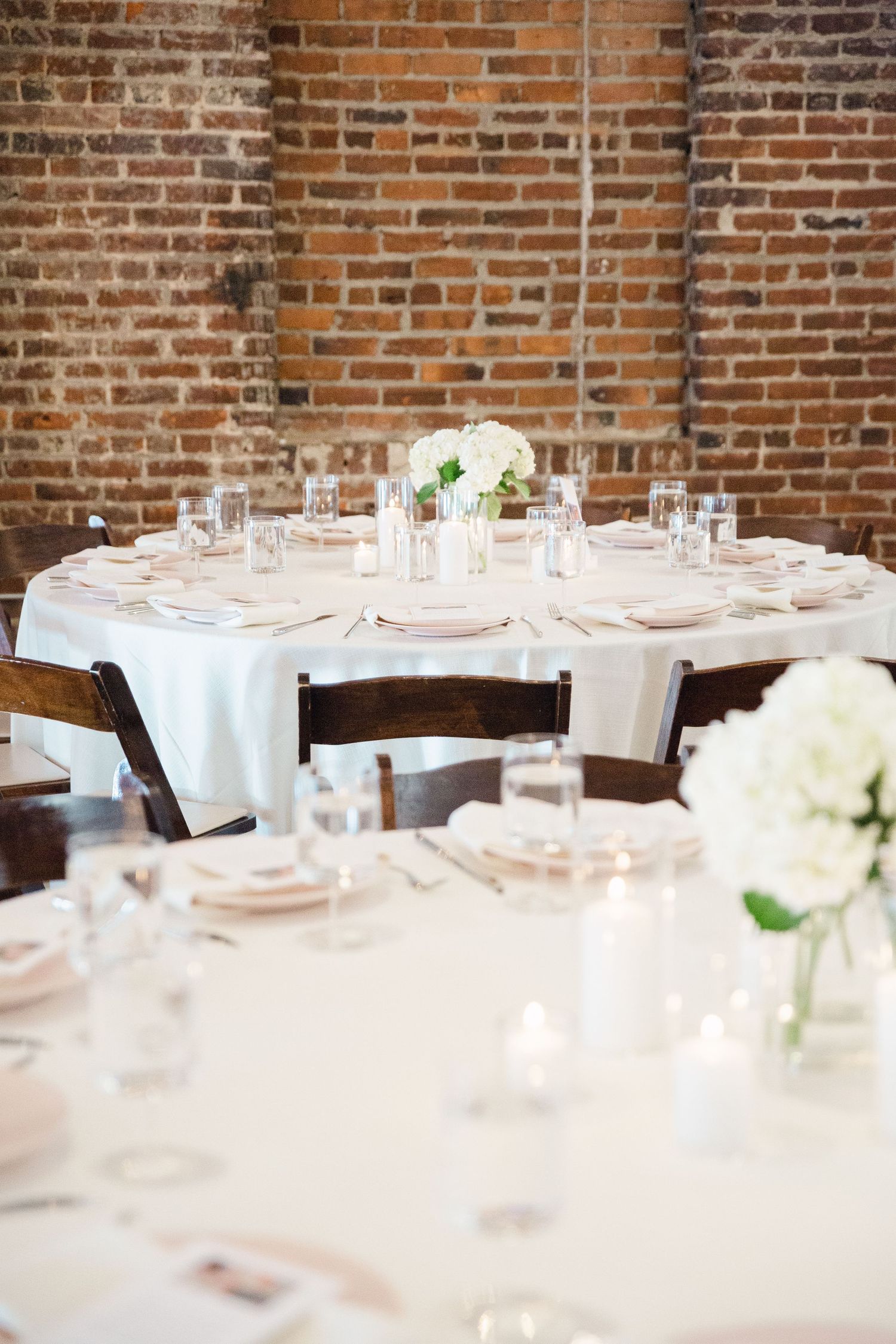 Round dining tables set with white linens and floral centerpieces in rustic brick venue.