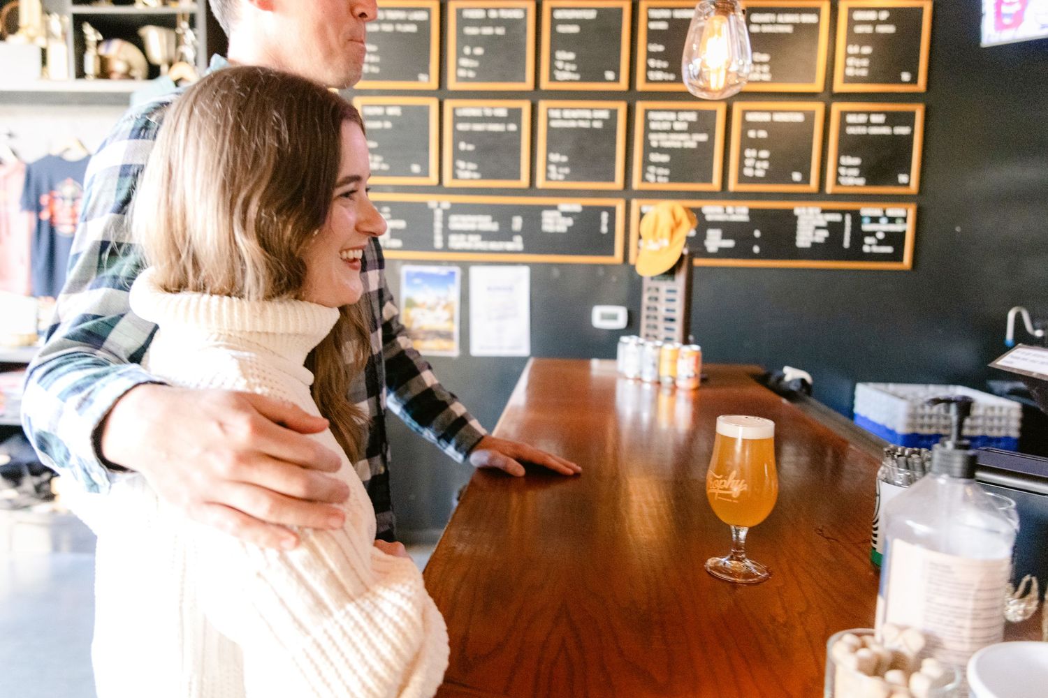 Two people share a warm embrace at a casual bar setting with wood furniture and a menu board on the wall.
