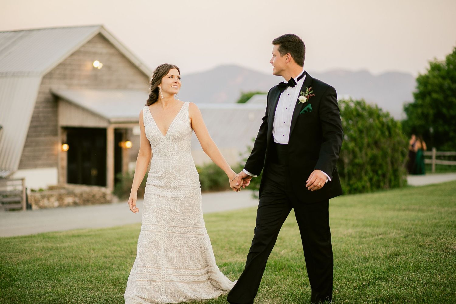 Couple walks hand in hand on grass with rustic barn and mountains in background.