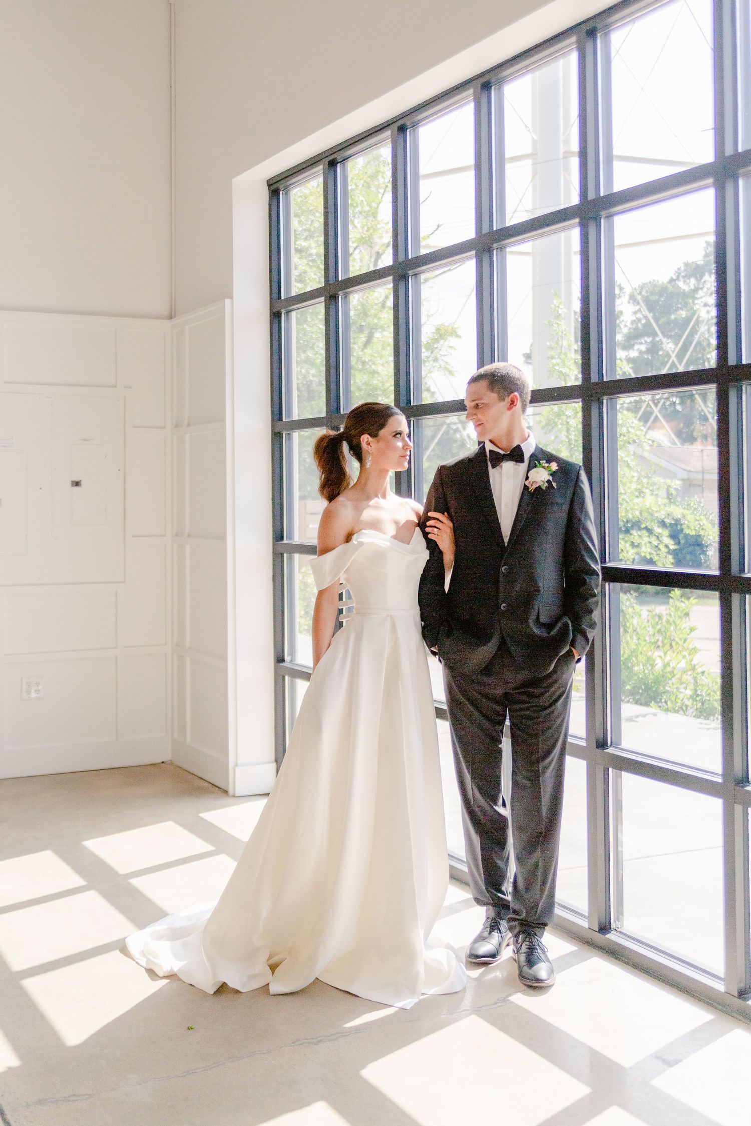 Wedding couple shares intimate moments in bright sunlit room with large windows casting dramatic shadows.