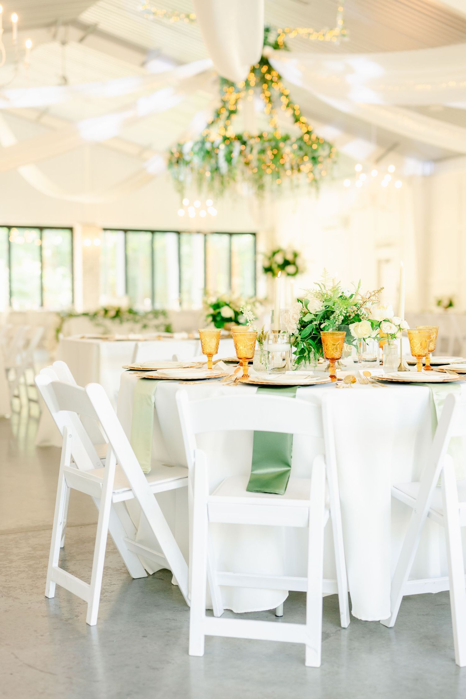 White folding chairs and tables set up in an elegant indoor wedding venue with greenery and floral decorations.