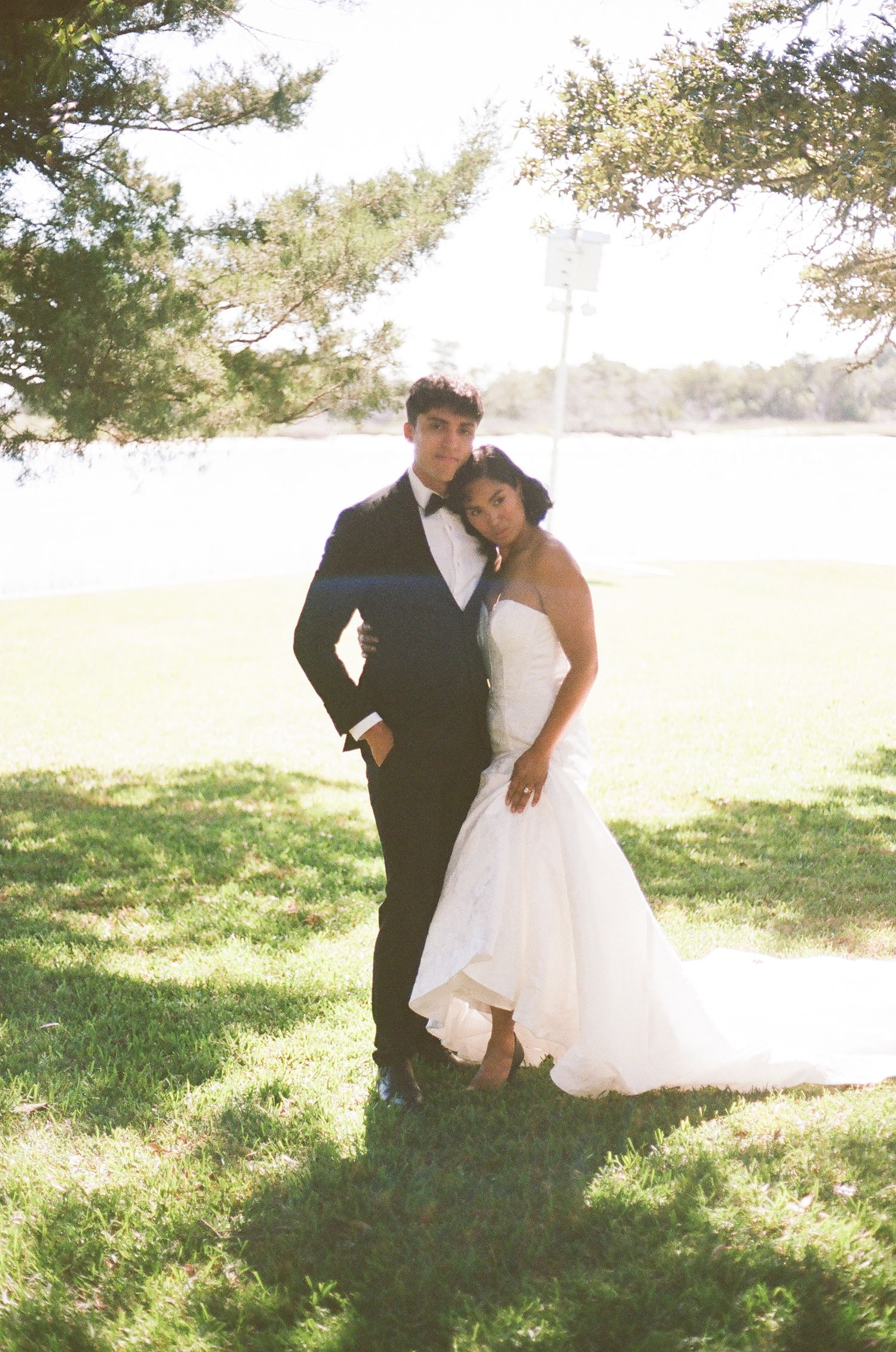 A couple in formal wedding attire poses together on a sunny grassy hillside.