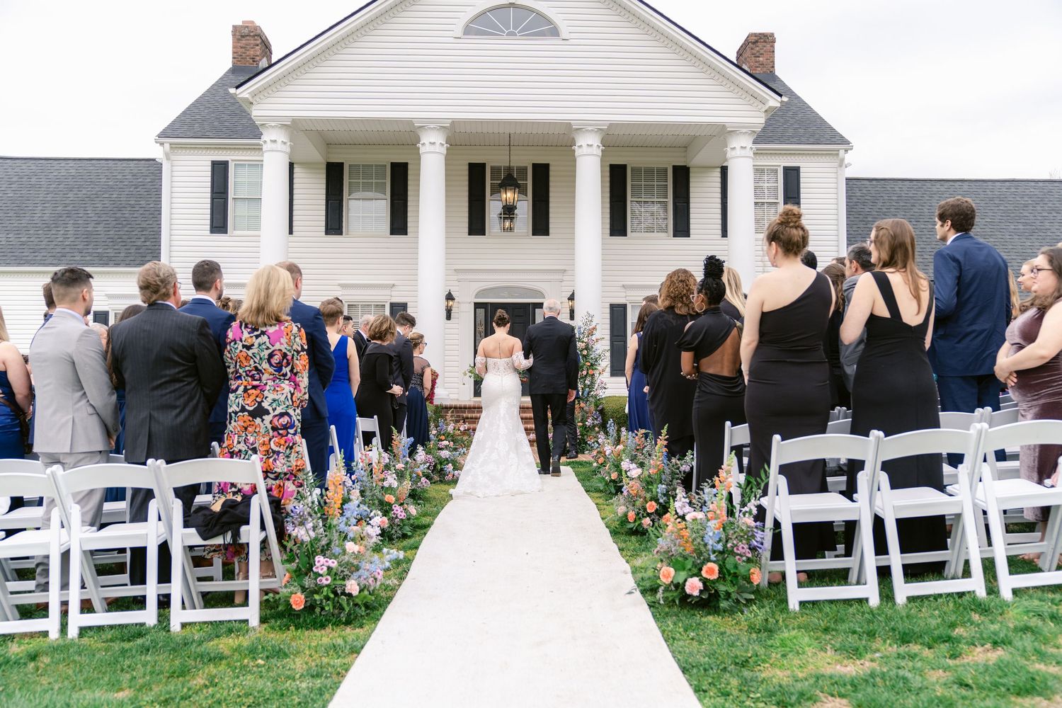 Outdoor wedding ceremony setup with white chairs on a lawn in front of a white colonial-style house with gray roof.