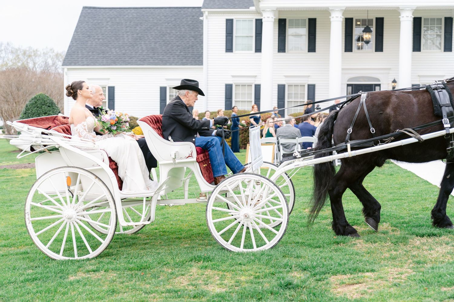 Elegant white horse-drawn carriage with a black horse stands on a manicured lawn in front of a colonial style house.