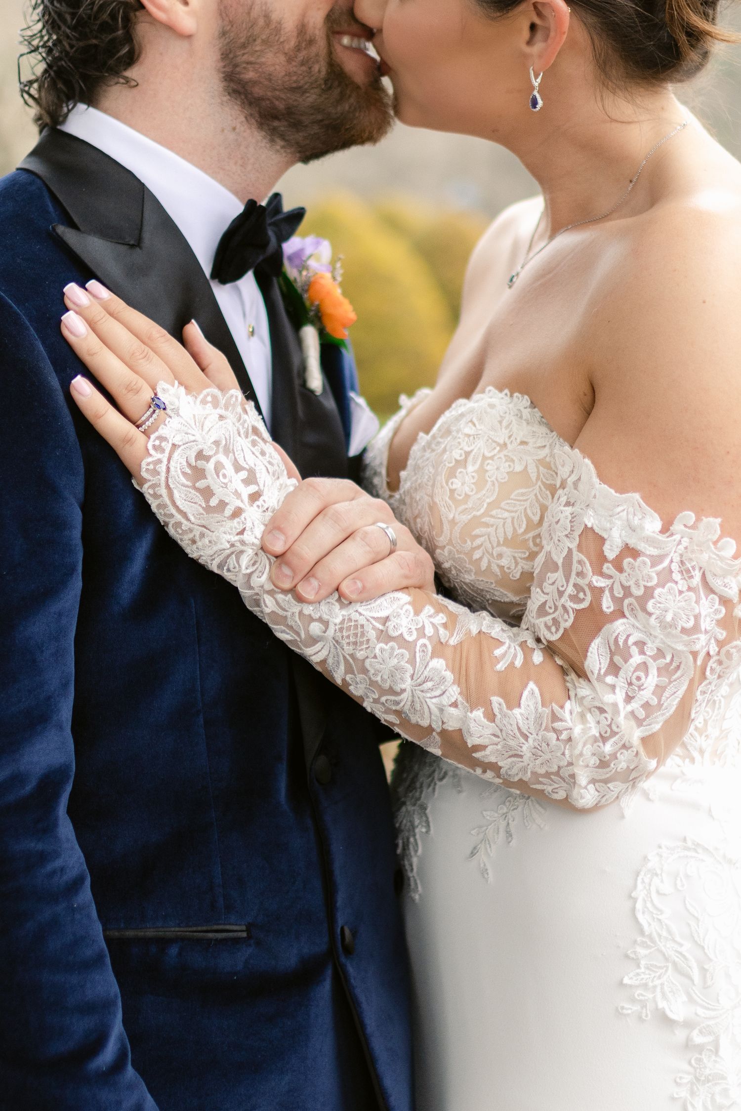A close-up wedding photo showing intricate lace sleeve details and a navy suit with a boutonniere.