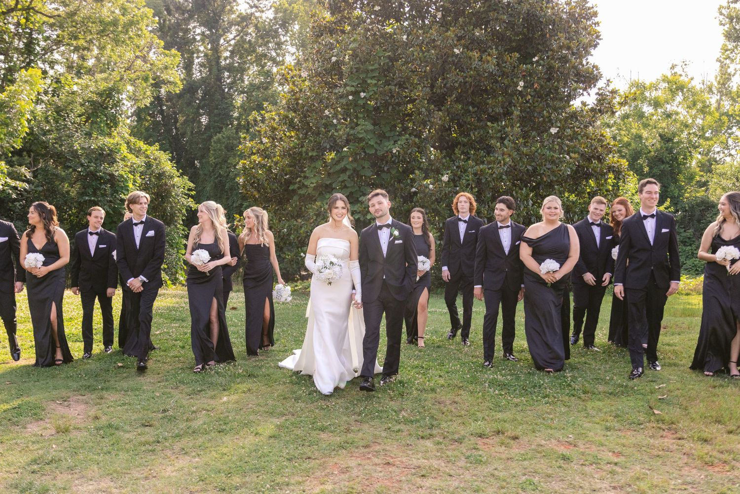 Wedding party in formal attire poses on grass with trees in background during outdoor photo session.