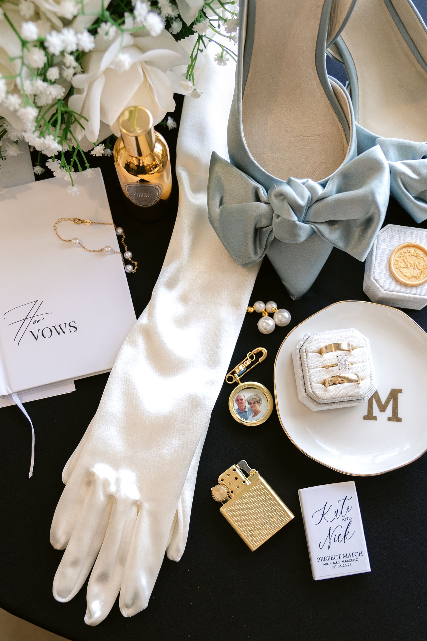 Elegant wedding accessories including blue bow heels, white gloves, jewelry, vows booklet, and decorative details on dark surface.