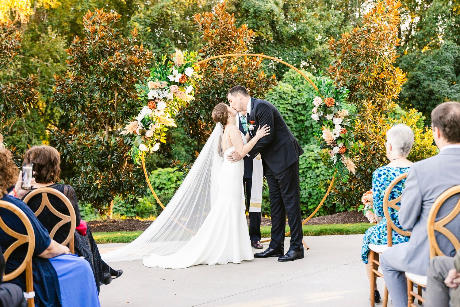Outdoor wedding ceremony in a garden with fall foliage backdrop and decorated floral arch.