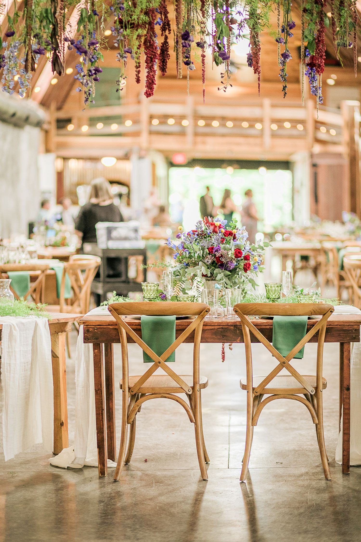 Elegant reception table setup with wooden chairs, green napkins, and hanging flower arrangements.