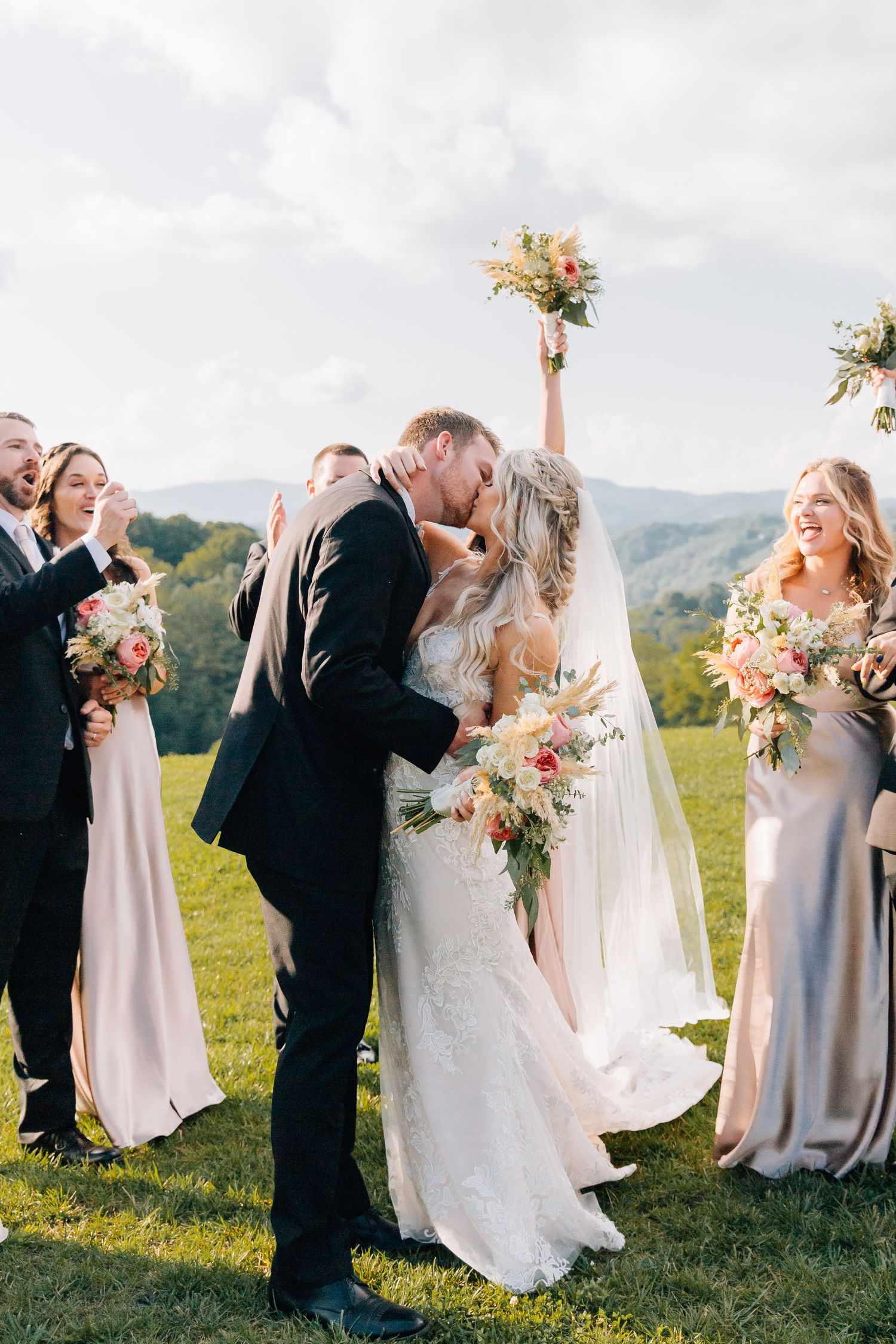 Wedding couple shares intimate moment surrounded by wedding party on scenic hilltop.
