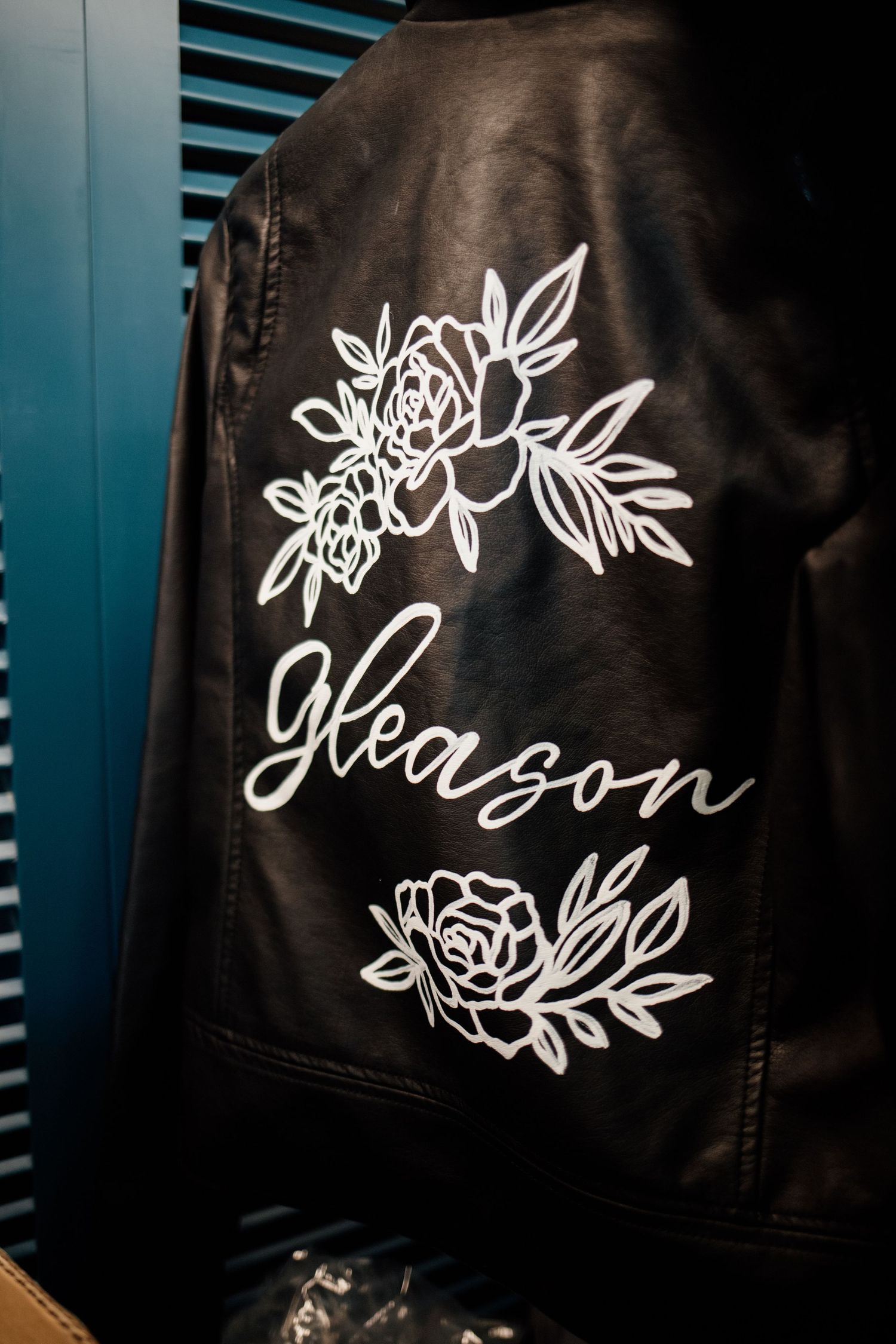 Black leather jacket with white floral embroidery and 'Gleason' script text displayed on the back.