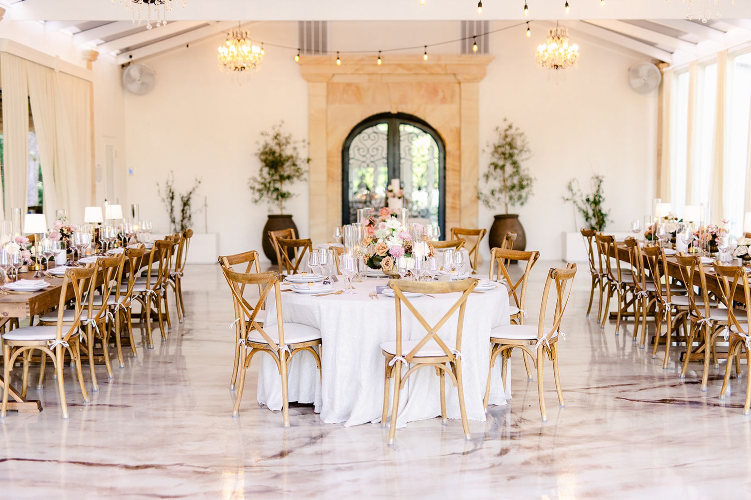 Elegant white wedding reception venue with rustic wooden cross-back chairs and white table settings in a bright space.