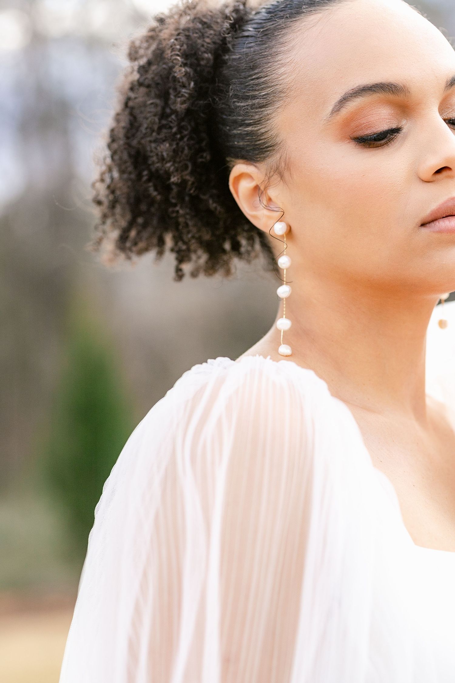 A woman with natural curly hair wearing pearl earrings and a light pink blouse poses against a soft blurred background.