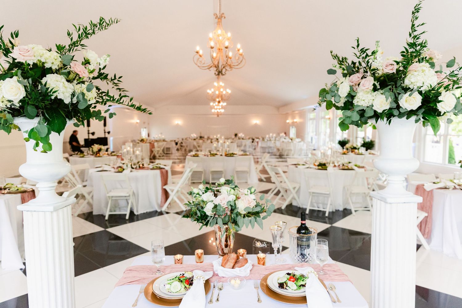 Elegant white wedding reception venue with crystal chandeliers, round tables, and tall floral centerpieces in white and green.