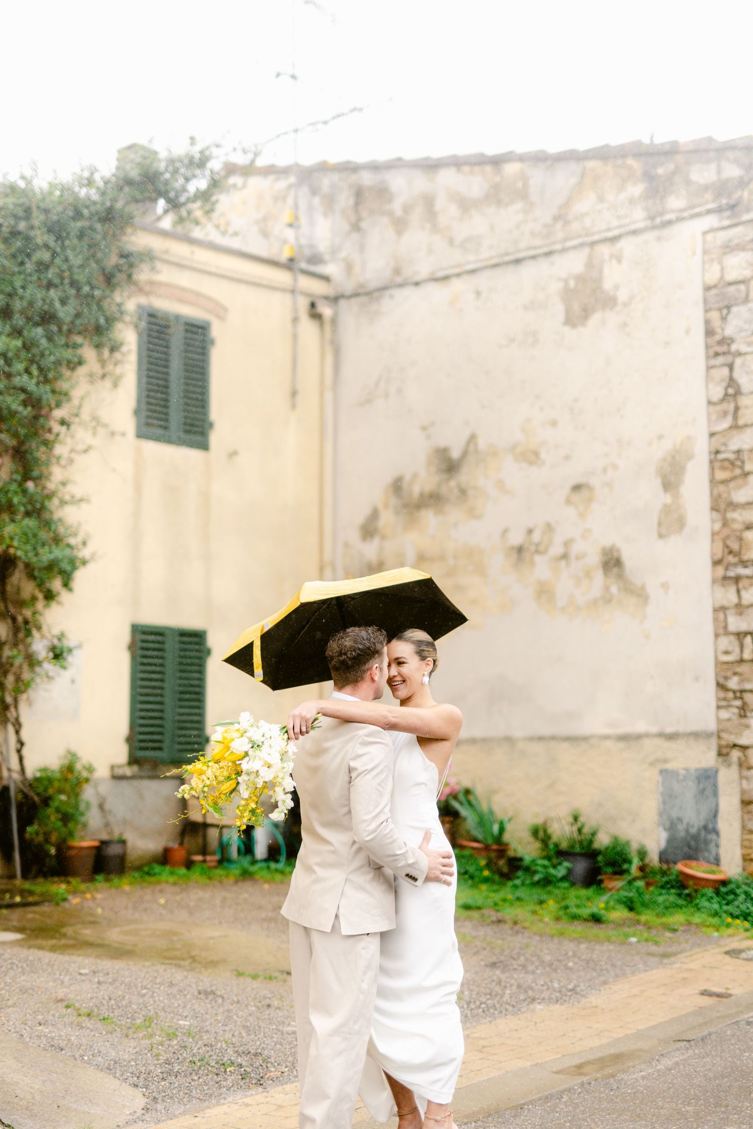 A couple in white outfits embraces and dances playfully against a rustic Mediterranean building backdrop.