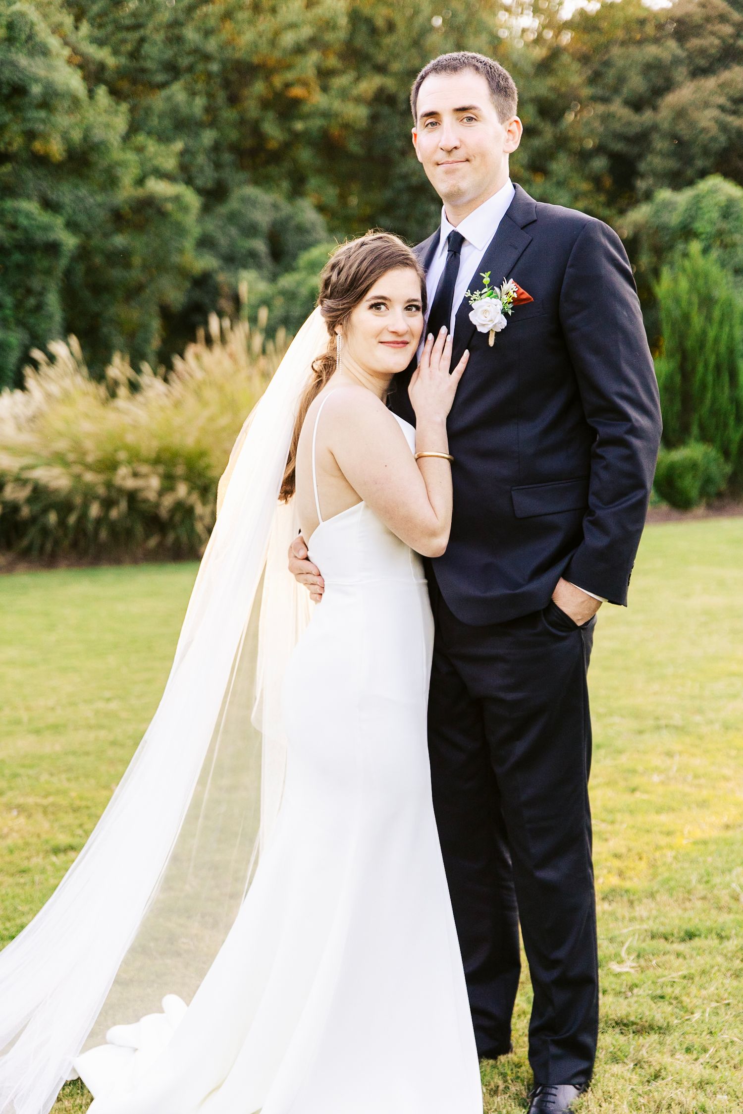 A bride and groom pose together outdoors on their wedding day in a garden setting with natural lighting.