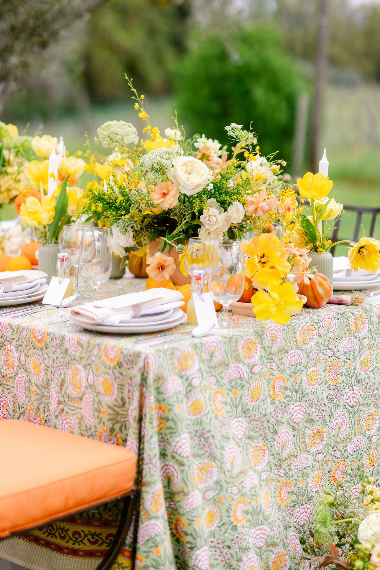 Elegant outdoor table setting with yellow and peach floral centerpiece on floral print tablecloth with orange cushioned chairs.