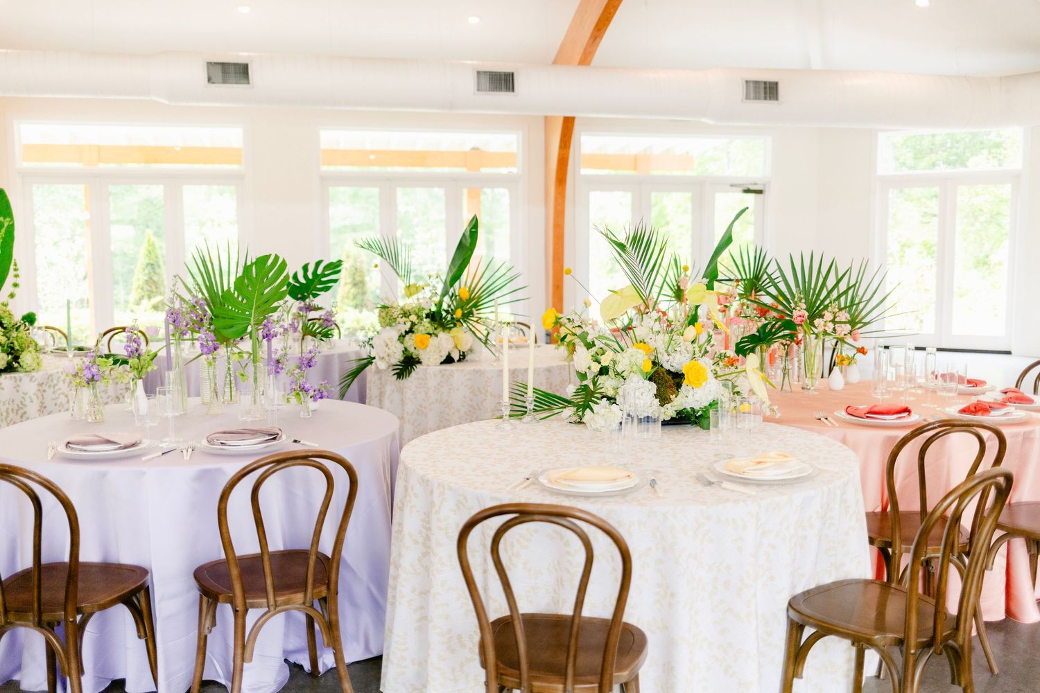 Light-filled reception venue with wooden chairs and white tablecloths set for wedding celebration.