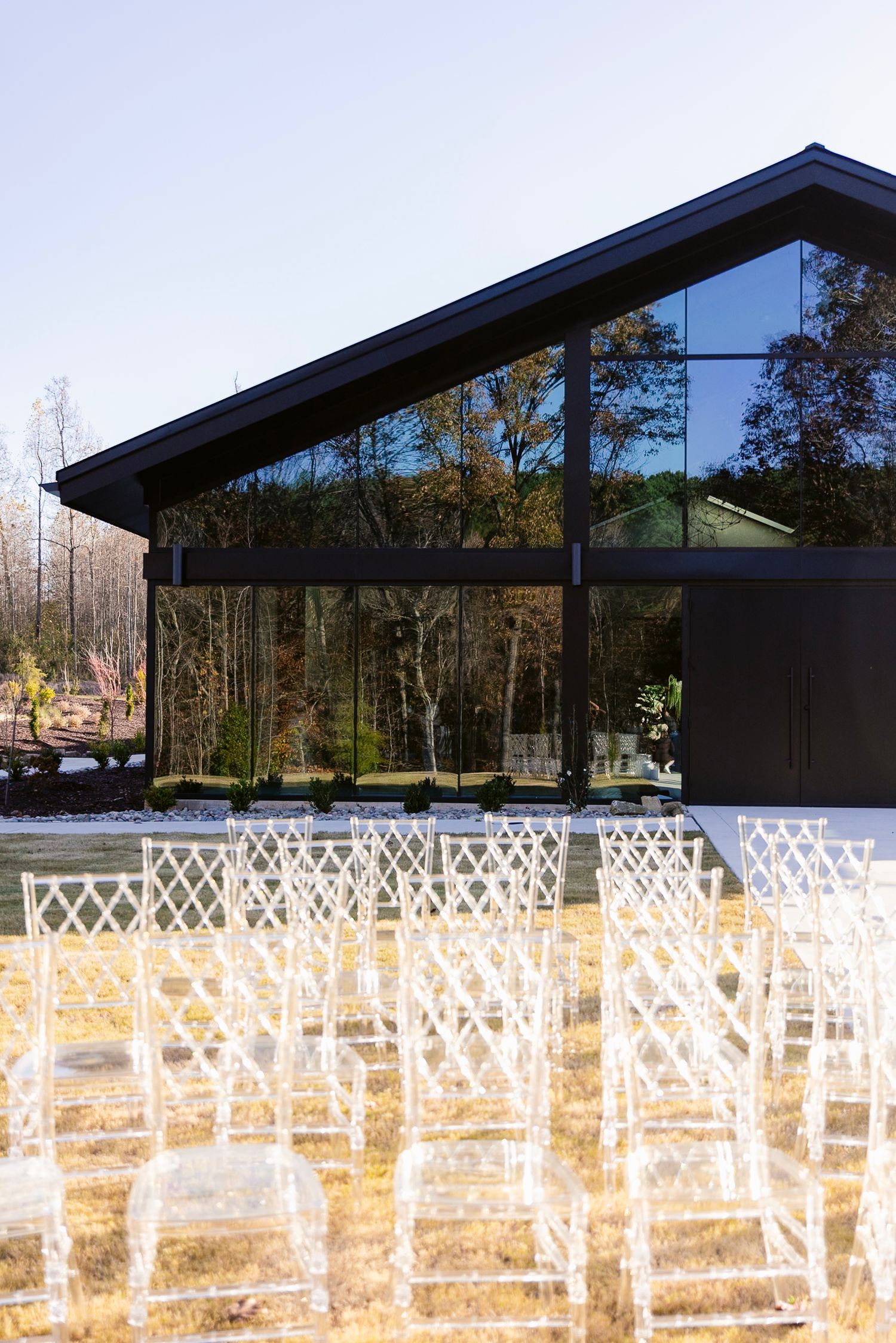 Modern glass pavilion with slanted roof and rows of clear acrylic chairs set up for outdoor event seating.