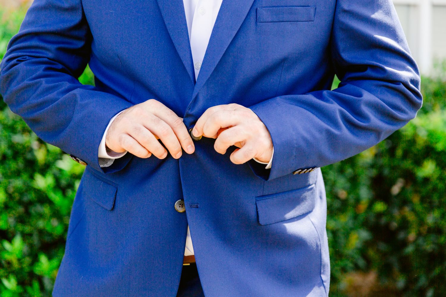 A person in a navy blue suit jacket stands outdoors against a green leafy background.