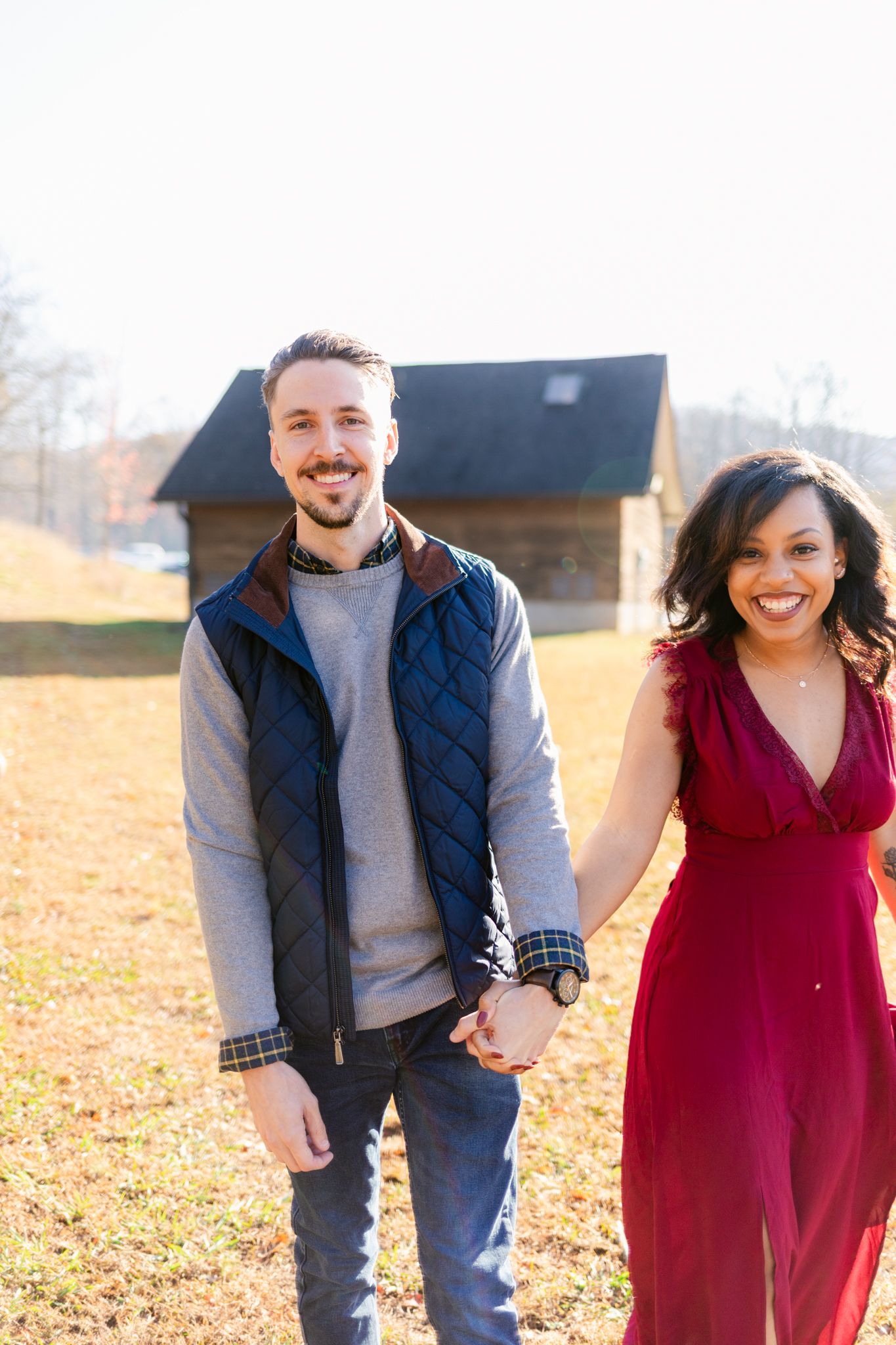 A couple in burgundy dress and navy vest laugh together while walking through a fall field on a sunny day.