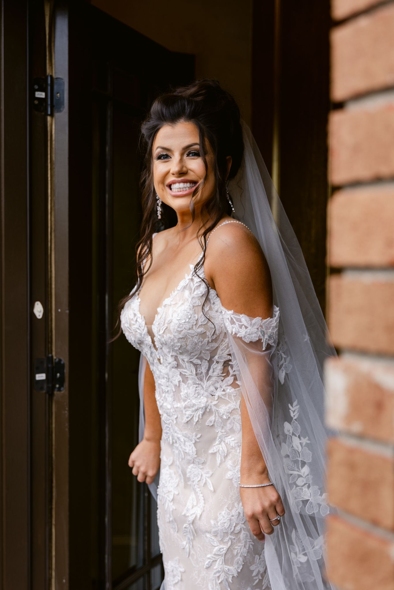 A bride in a beautiful white lace wedding dress with off-shoulder details poses against a brick wall background.