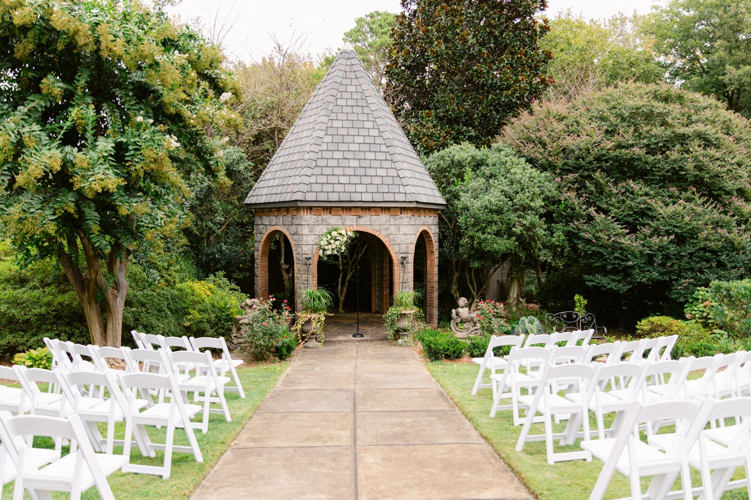 White wedding chairs line a garden pathway leading to a wooden gazebo surrounded by lush greenery and trees.