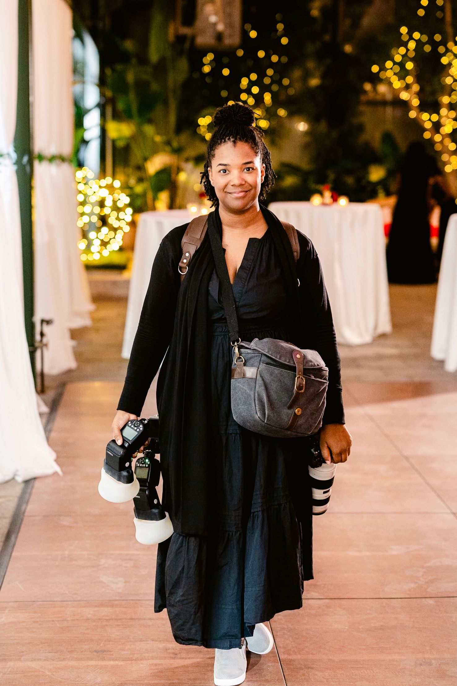 A person in a black blazer and dress walks through an outdoor plaza with twinkling lights in the background.