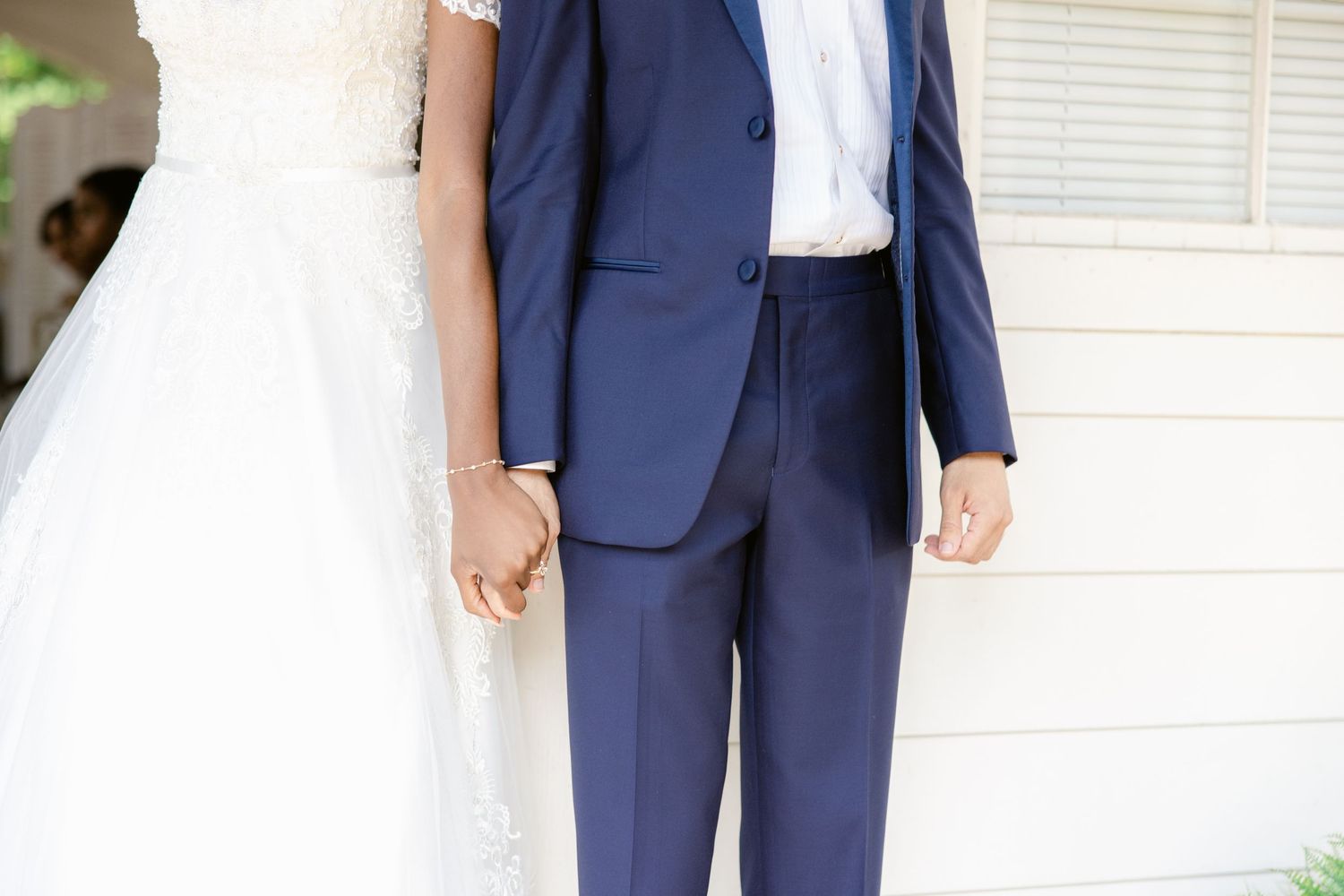 A couple in wedding attire stands together on a porch with white columns and green plants in the background.