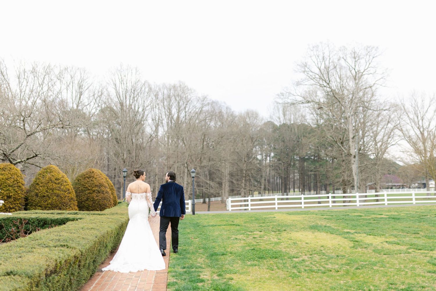 Newlywed couple walks along a scenic country path surrounded by bare winter trees and green fields.