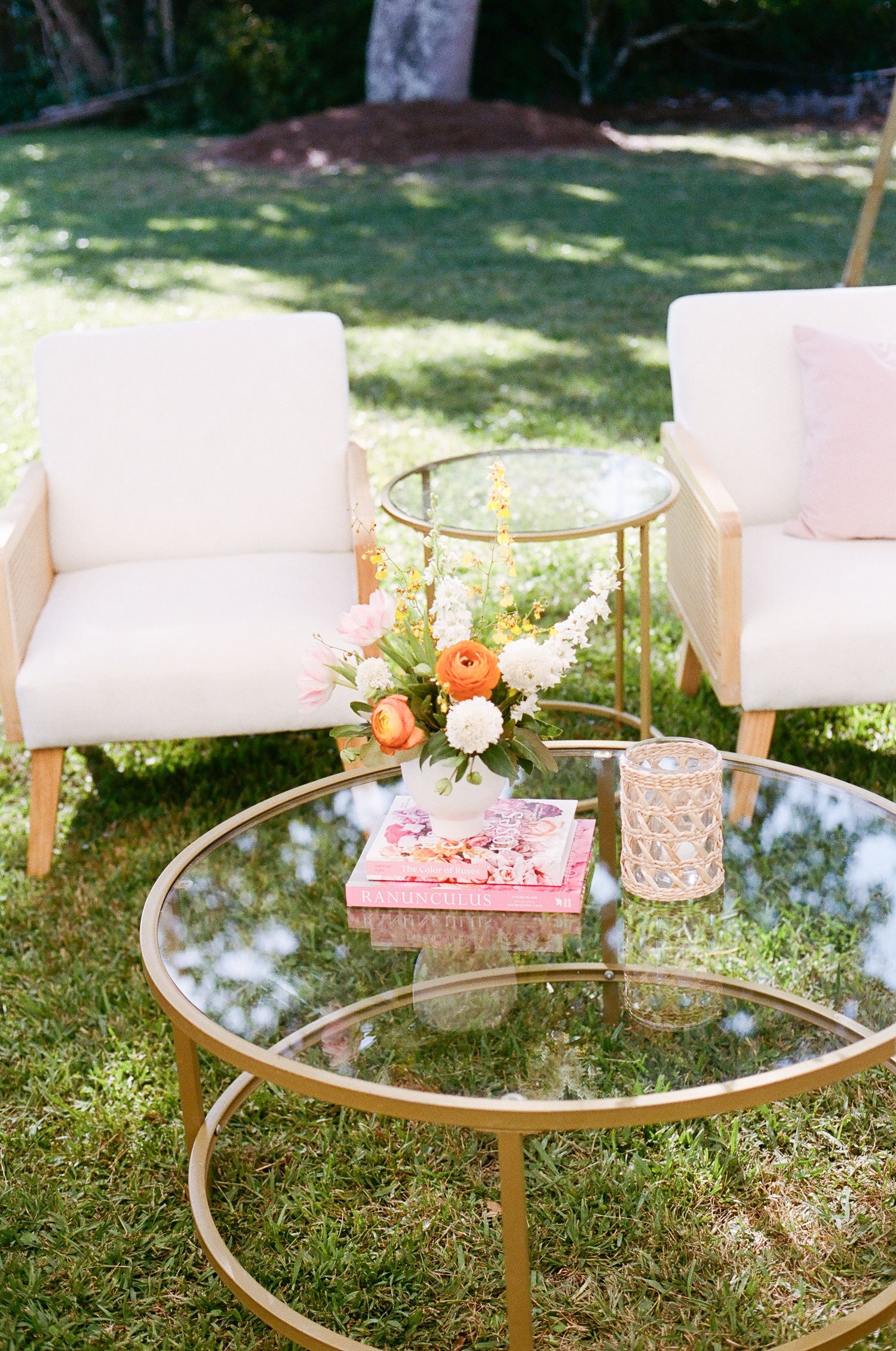 Round glass coffee tables with gold frames arranged on grass with white cushioned seating for an outdoor gathering.